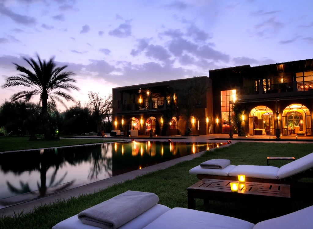 Resort terrace at dusk: a palm tree beside a warmly lit building with arched windows and a reflecting pool, white lounge chairs in the foreground.